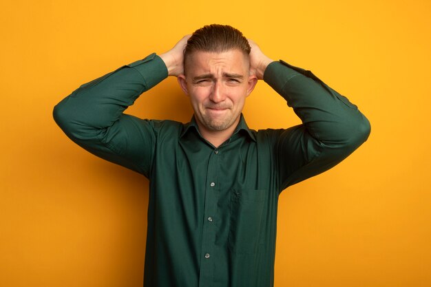 Hombre guapo joven en camisa verde mirando al frente confundido con expresión molesta tomados de la mano detrás de la cabeza de pie sobre la pared naranja