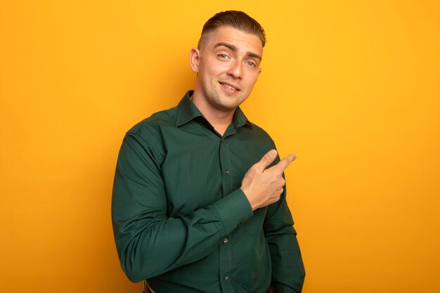 Hombre guapo joven en camisa verde sonriendo apuntando con el dedo índice hacia atrás