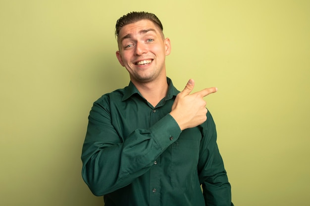 Hombre guapo joven en camisa verde con una sonrisa en la cara apuntando con el dedo índice hacia el lado