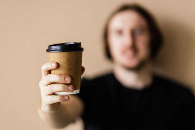 Hombre guapo joven con camiseta beige y anteojos sosteniendo una taza de café para llevar en una mano satisfecho con el tiempo libre sobre fondo beige
