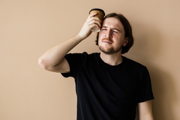 Hombre guapo joven con camiseta beige y anteojos sosteniendo una taza de café para llevar en una mano satisfecho con el tiempo libre sobre fondo beige