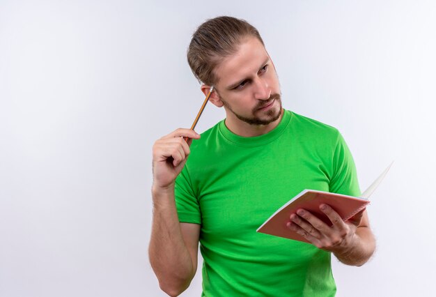 Hombre guapo joven en camiseta verde con cuaderno y lápiz mirando a un lado con expresión pensativa en la cara de pie sobre fondo blanco.