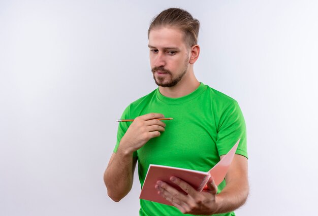 Hombre guapo joven en camiseta verde con cuaderno y lápiz mirando a un lado con expresión pensativa en la cara de pie sobre fondo blanco.