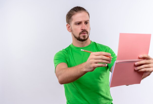 Hombre guapo joven en camiseta verde con cuaderno y lápiz mirando portátil con expresión pensativa en la cara de pie sobre fondo blanco.