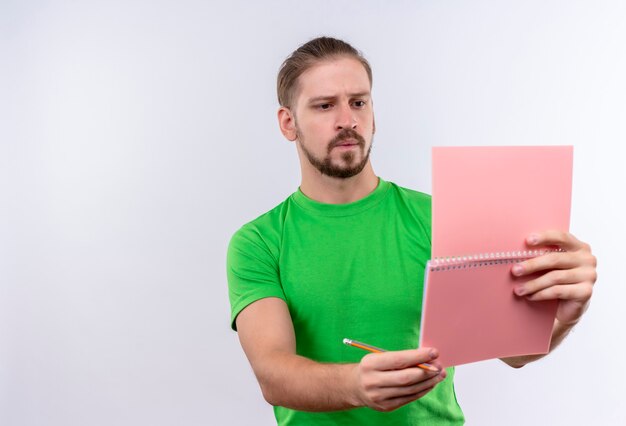 Hombre guapo joven en camiseta verde con cuaderno y lápiz mirando portátil con expresión pensativa en la cara de pie sobre fondo blanco.