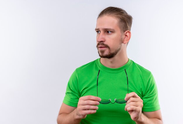 Hombre guapo joven en camiseta verde con gafas en las manos mirando a un lado con seria expresión de confianza de pie sobre fondo blanco