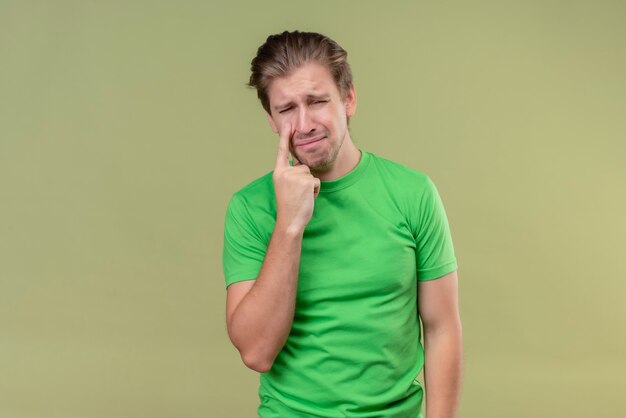 Hombre guapo joven con camiseta verde llorando con expresión triste en la cara de pie sobre la pared verde