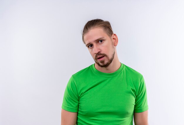 Hombre guapo joven en camiseta verde mirando confundido y muy ansioso de pie sobre fondo blanco.
