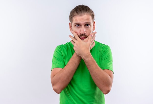 Hombre guapo joven en camiseta verde mirando confundido y sorprendido sosteniendo su rostro con las manos de pie sobre fondo blanco.