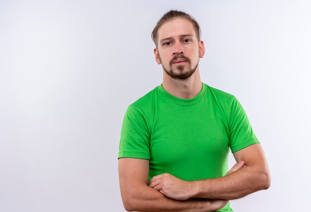 Hombre guapo joven en camiseta verde de pie con los brazos cruzados mirando a la cámara con cara seria sobre fondo blanco.