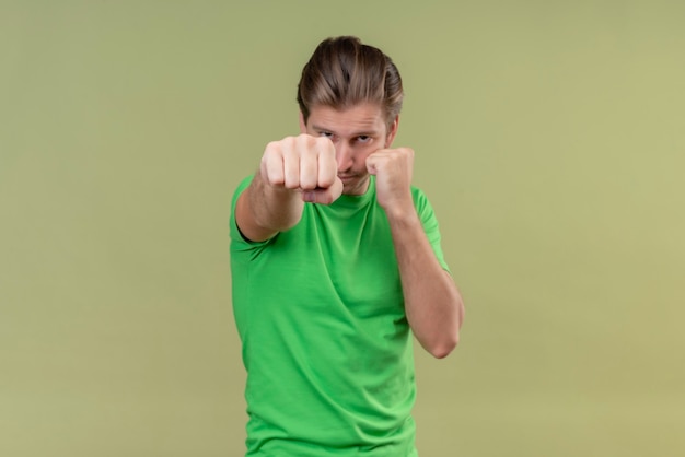 Hombre guapo joven con camiseta verde posando como un boxeador con el puño cerrado de pie sobre la pared verde