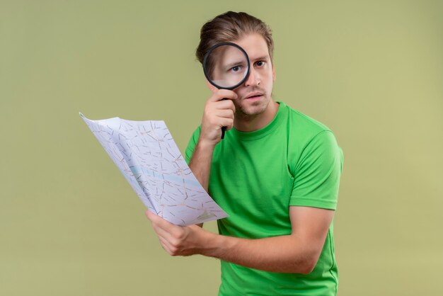 Hombre guapo joven con camiseta verde sosteniendo el mapa mirando a través de una lupa a la cámara con expresión seria en la cara de pie sobre la pared verde