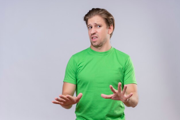Hombre guapo joven con camiseta verde sosteniendo sus manos en alto diciendo que no te acerques con expresión de disgusto de pie sobre la pared blanca
