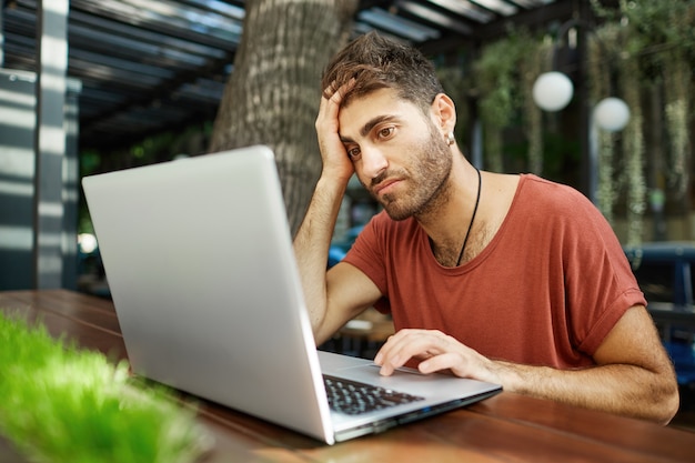 Hombre guapo joven cansado sentado con la computadora portátil en la cafetería al aire libre, trabajando de forma remota o estudiando con wifi del parque