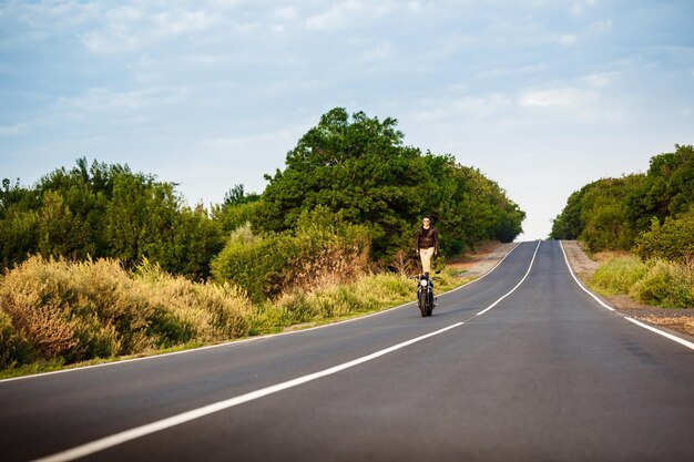 Hombre guapo joven con chaqueta de cuero montando en moto, haciendo trucos en la carretera del campo.