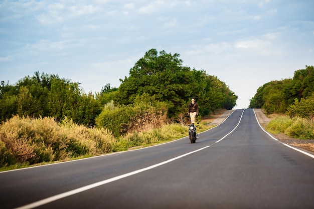 Hombre guapo joven con chaqueta de cuero montando en moto, haciendo trucos en la carretera del campo.