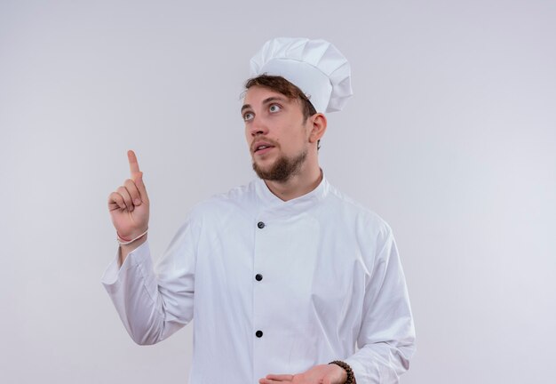 Un hombre guapo joven chef barbudo vestido con uniforme de cocina blanco y sombrero apuntando hacia arriba con el dedo índice sobre una pared blanca