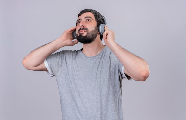 Hombre guapo joven complacido con auriculares escuchando música y mirando hacia arriba con las manos en los auriculares aislados en la pared blanca