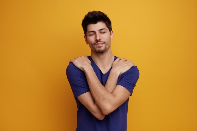 Hombre guapo joven complacido manteniendo las manos sobre los hombros con los ojos cerrados sobre fondo amarillo