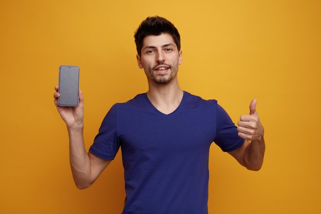 Hombre guapo joven complacido mirando a la cámara mostrando el teléfono móvil a la cámara y mostrando el pulgar hacia arriba sobre fondo amarillo