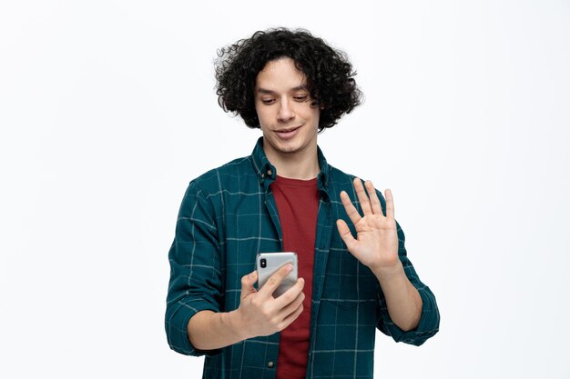 Hombre guapo joven complacido sosteniendo teléfono móvil hablando a través de videollamadas agitando aislado sobre fondo blanco.