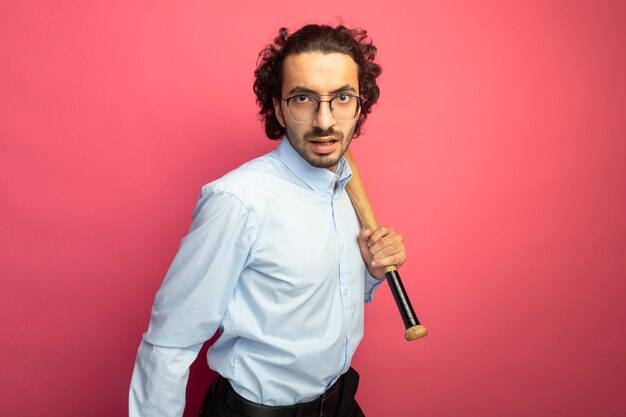 Hombre guapo joven confiado con gafas sosteniendo un bate de béisbol mirando al frente aislado en la pared rosa