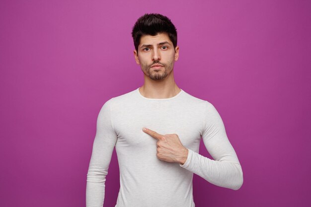 Hombre guapo joven confiado mirando a la cámara apuntándose a sí mismo sobre fondo púrpura