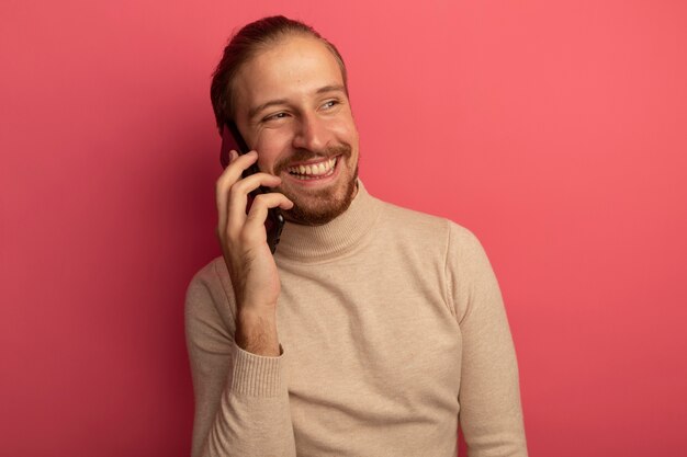 Hombre guapo joven en cuello alto beige sonriendo alegremente mientras habla por teléfono móvil de pie sobre la pared rosa