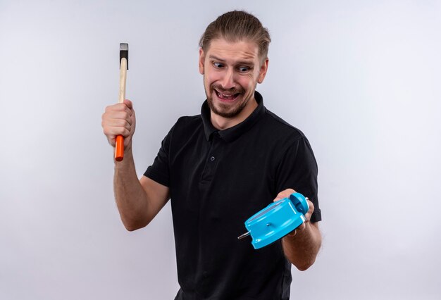 Hombre guapo joven decepcionado en camisa polo negra sosteniendo el reloj de alarma y balanceando un martillo que va a romper el reloj de pie sobre fondo blanco.