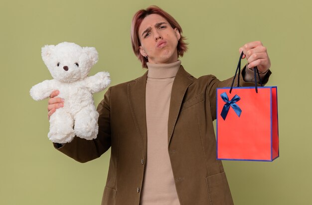 Hombre guapo joven disgustado con oso de peluche blanco y bolsa de regalo