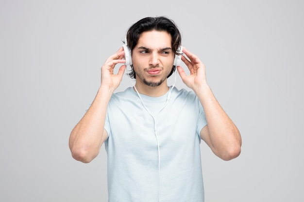 Hombre guapo joven disgustado usando y tocando auriculares mirando al lado aislado sobre fondo blanco.