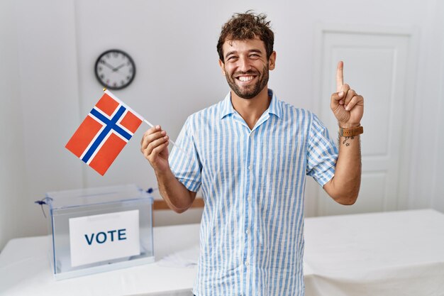 Hombre guapo joven en la elección de la campaña política con la bandera de noruega sonriendo con una idea o pregunta señalando con el dedo con la cara feliz número uno