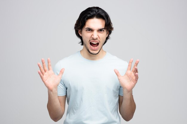 Hombre guapo joven enojado manteniendo las manos en el aire mirando a la cámara gritando aislado sobre fondo blanco.