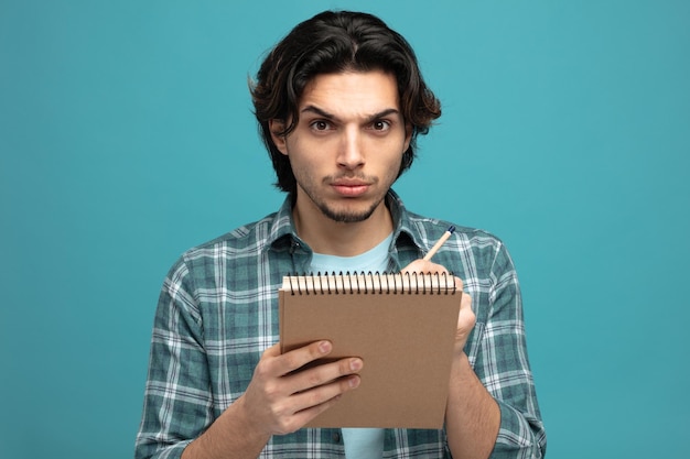 hombre guapo joven estricto sosteniendo un bloc de notas y un lápiz mirando a la cámara aislada en el fondo azul