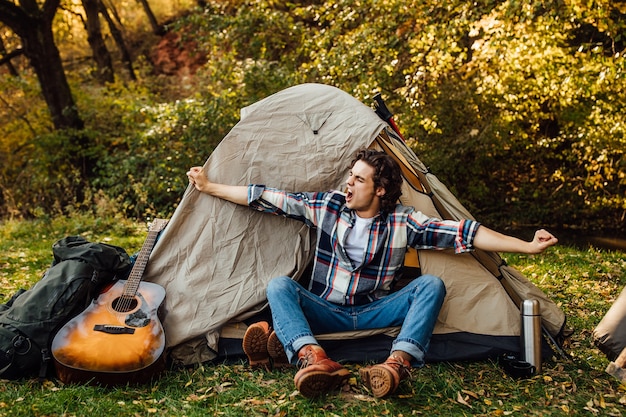 Hombre guapo joven se extiende por la mañana cerca de la tienda de campaña en el camping en la naturaleza