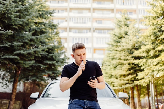 Hombre guapo joven fumando con vape cerca del vehículo