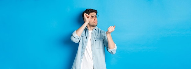 Hombre guapo joven en gafas mirando sus uñas comprobando la manicura de pie sobre backgr azul