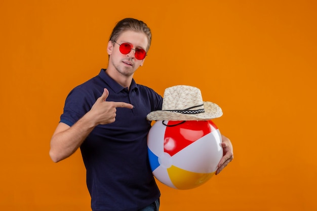 Hombre guapo joven con gafas de sol rojas sosteniendo una pelota inflable y un sombrero de paja de verano apuntando con el dedo mirando a la cámara con expresión de confianza en la cara de pie sobre fondo naranja