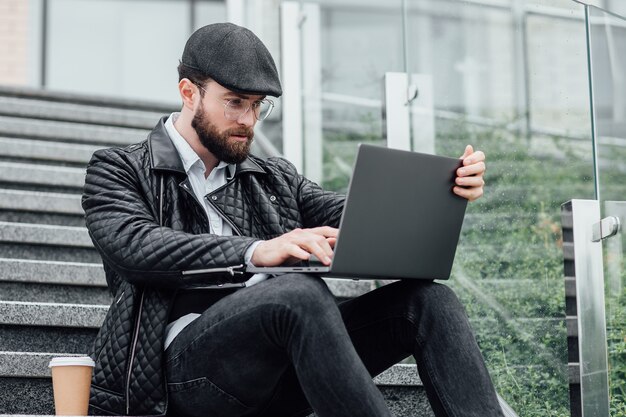 Hombre guapo joven gerente tomando café y trabajando en la computadora portátil mientras está sentado al aire libre en las escaleras