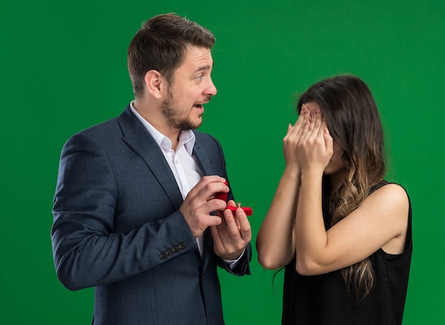 Hombre guapo joven hermosa pareja va a hacer una propuesta a su encantadora novia emocionada con los ojos cerrados celebrando el día de San Valentín de pie sobre la pared verde