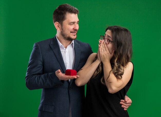 Hombre guapo joven hermosa pareja sosteniendo una caja roja con anillo de compromiso va a hacer una propuesta a su encantadora novia emocionada celebrando el día de San Valentín de pie sobre la pared verde
