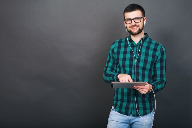 Hombre guapo joven inconformista sobre fondo gris con tableta escuchando música en auriculares hablando en línea feliz sonriendo anteojos de camisa a cuadros verde, estado de ánimo positivo