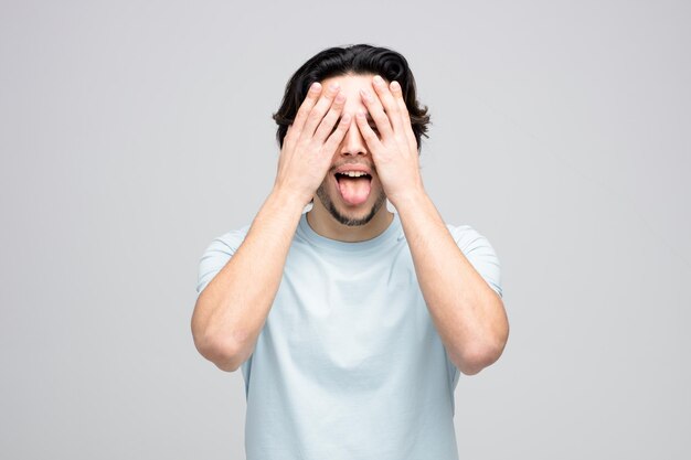 Hombre guapo joven juguetón cerrando los ojos con las manos mostrando la lengua aislado sobre fondo blanco.