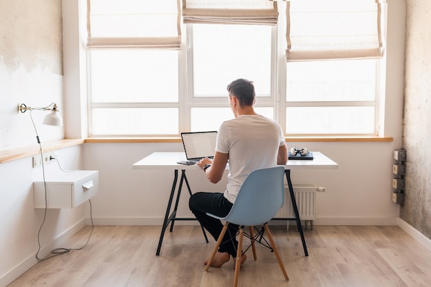 Hombre guapo joven moderno en traje casual sentado en la mesa trabajando en la computadora portátil, autónomo en casa