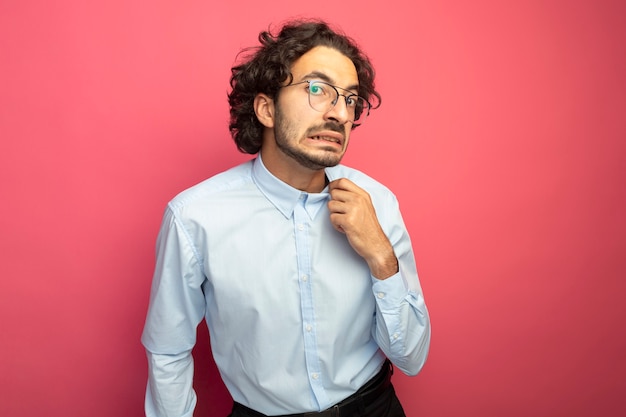 Hombre guapo joven molesto con gafas mirando al frente agarrando el cuello de la camisa aislado en la pared rosa