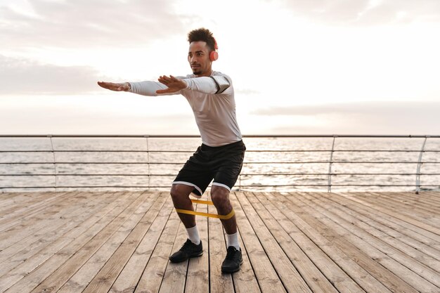 Hombre guapo joven en pantalones cortos negros y camiseta blanca escucha música en auriculares y se pone en cuclillas con goma de fitness cerca del mar