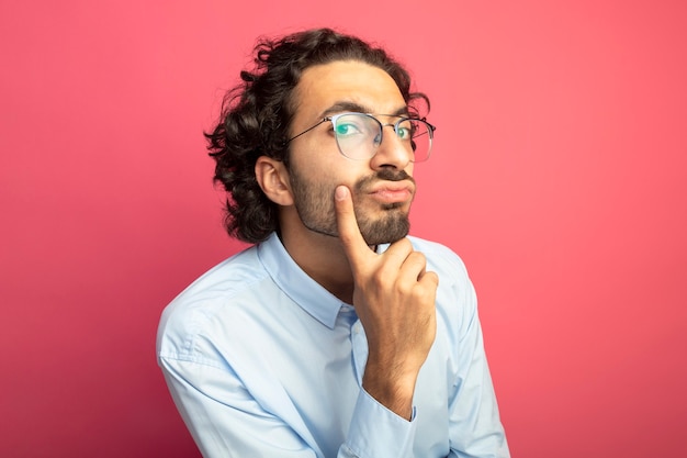 Hombre guapo joven pensativo con gafas mirando al frente tocando la barbilla aislada en la pared rosa