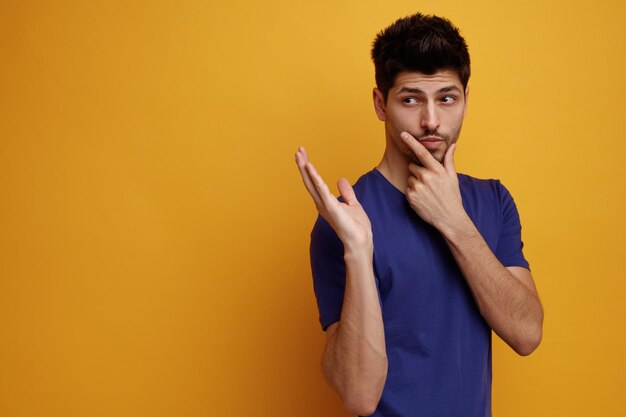 Hombre guapo joven pensativo manteniendo la mano en la barbilla mostrando la mano vacía mirando el lado sobre fondo amarillo con espacio de copia