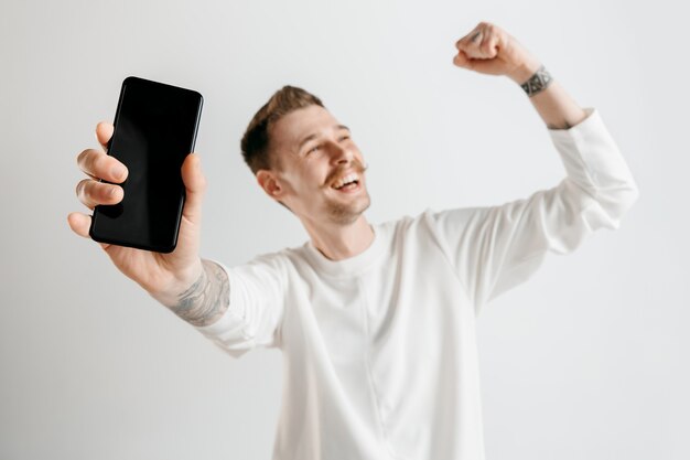 Hombre guapo joven que muestra la pantalla del teléfono inteligente en gris con una cara de sorpresa.