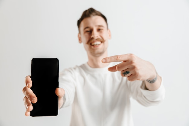 Hombre guapo joven que muestra la pantalla del teléfono inteligente en gris con una cara de sorpresa.